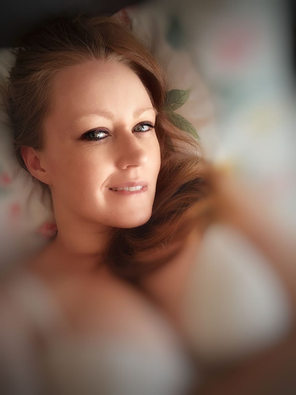 An photo of Sin looking directly into the camera as she bites her lip. She's lying in bed, wearing a white bra.